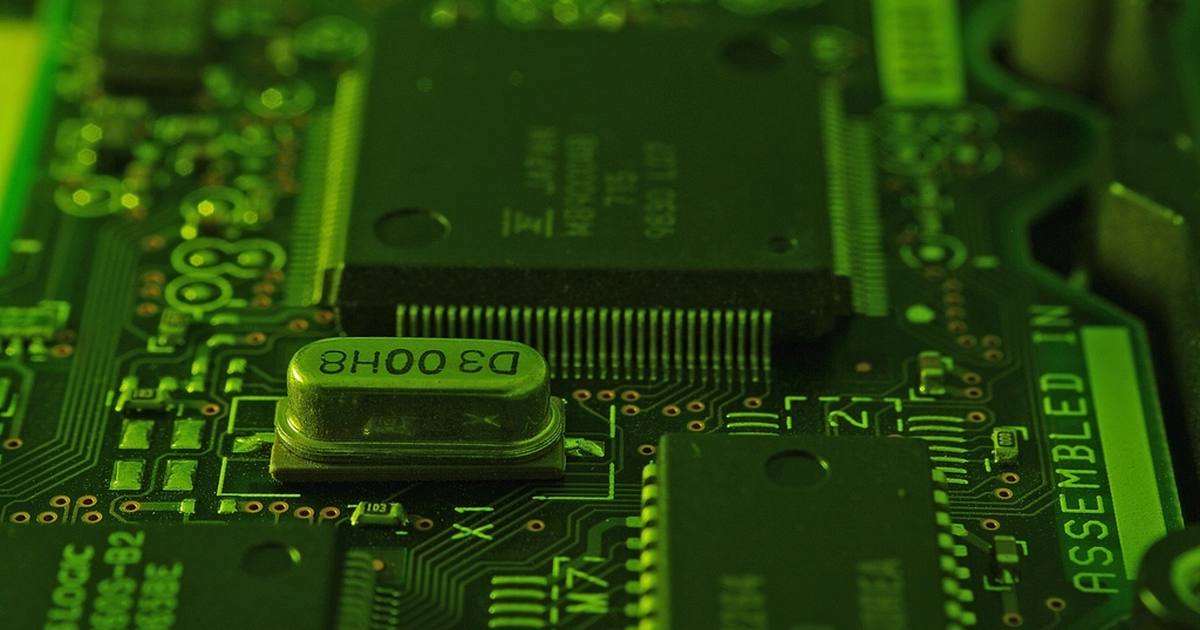 Close-up of a green-lit circuit board with a large surface-mount chip, a crystal oscillator and fine copper traces