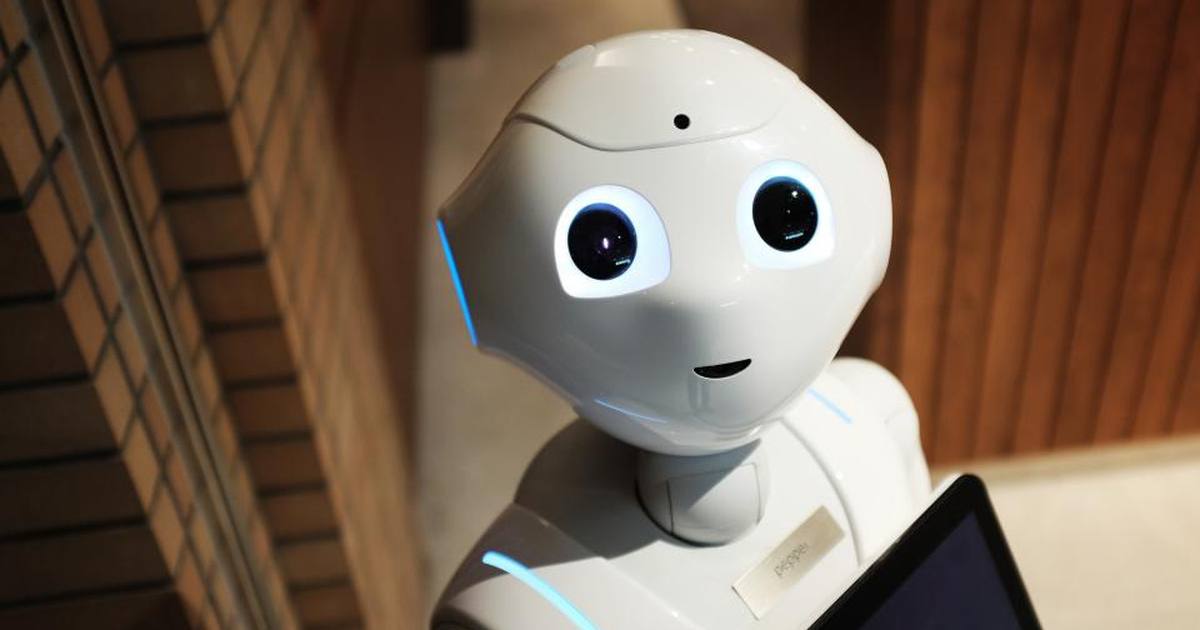 A white humanoid service robot with large dark eyes, holding a tablet in front of a brick and wood-panelled wall
