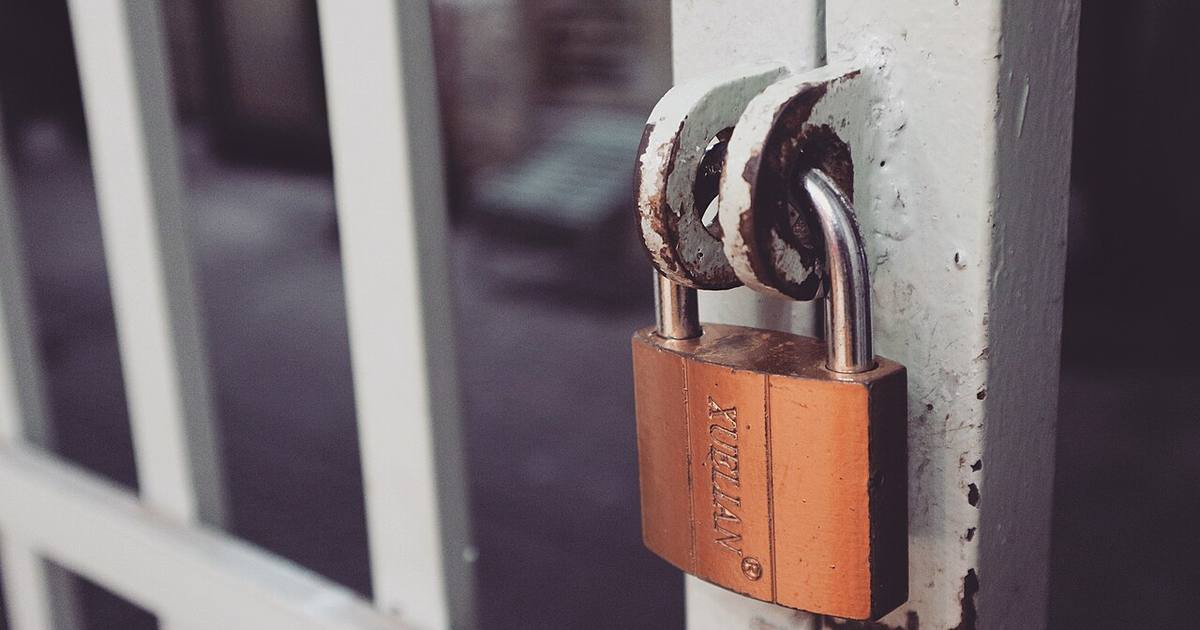 A worn brass padlock fastened through the hasp of a white-painted metal gate